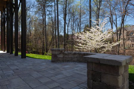 Malerischer Blick auf den Hinterhof im Frühling mit Terrassenpflaster und Steinmauer, blühenden weißen Kirschbaum und frühlingshaften Wäldern im Hintergrund.