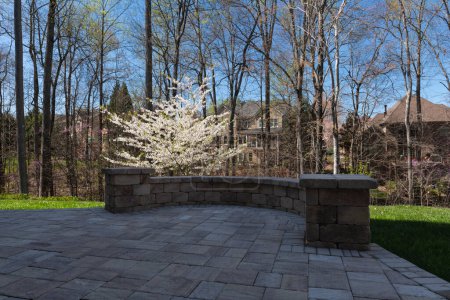 Malerischer Blick auf den Hinterhof im Frühling mit Terrassenpflaster und Steinmauer, blühenden weißen Kirschbaum und frühlingshaften Wäldern im Hintergrund.