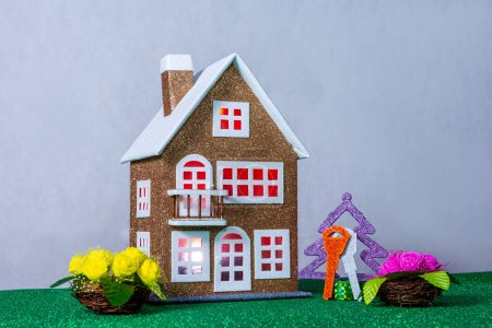 Cerca de una casa de juguete marrón con luz roja en las ventanas hay flores de colores y un árbol de Navidad con llaves
