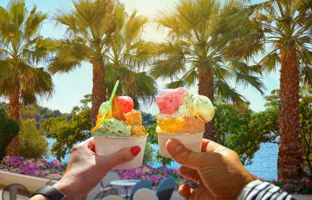 Foto de Couple with beautiful bright  sweet  ice-cream with different flavors  in the hands. Background of   view of the see with coconut palm trees in Porec ,Croatia.Traveling concept background - Imagen libre de derechos
