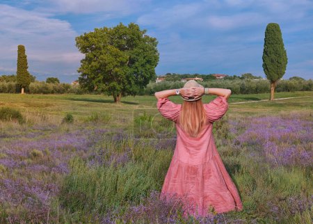 Photo for Back view of happy woman with hands up in straw hat  on a lavender field. Landscape with a cypress tree near the Green lagoon sea bay in Porec, Croatia - Istria, Europe - Royalty Free Image