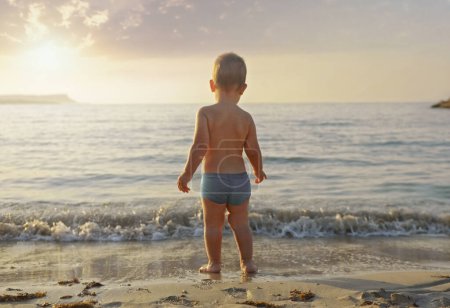 Foto de Un niño se para en la playa y mira al mar.Vacaciones con niños.Feliz concepto de estilo de vida infantil.Vista desde atrás - Imagen libre de derechos