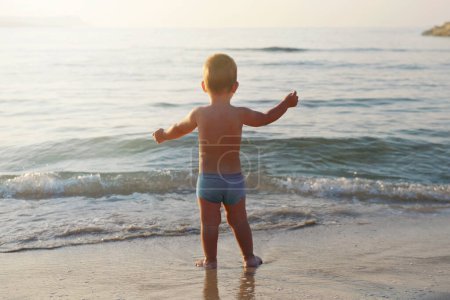 Téléchargez les photos : Un garçon se tient sur la plage et regarde la mer.Vacances avec des enfants.Concept d'enfance de mode de vie heureux.Vue de dos - en image libre de droit