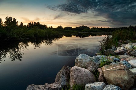 Téléchargez les photos : Paysage panoramique vue du magnifique coucher de soleil sur un lac pittoresque avec ciel et nuages réfléchis sur l'eau avec des roches et des pierres au premier plan. - en image libre de droit