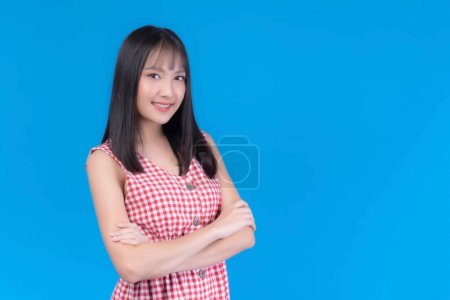 Foto de Retrato de hermosa mujer asiática linda chica en vestido rojo con flequillo estilo de pelo, risa y sonriente buen humor de pie brazos cruzados aislado fondo azul - - Imagen libre de derechos