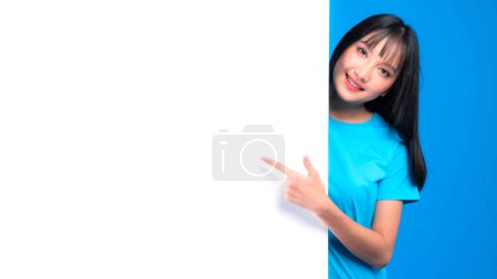 Photo for Beautiful Asian young woman with bangs hair style in blue t shirt smiling and pointing finger a blank space for advertising banner , empty space white board a blank banner isolated on blue background - Royalty Free Image