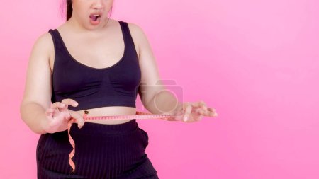 Téléchargez les photos : Asiatique grosses femmes, Fat girl, Joufflu, en surpoids serrer la graisse du ventre mesure avec du ruban à mesurer sur sa taille isolé sur fond rose - Femme alimentation mode de vie problème d'embonpoint concept - en image libre de droit