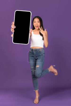 Téléchargez les photos : Full body length photo portrait of beautiful Asian young woman ,Excited surprised girl showing big smart phone with blank screen , white screen isolated on purple background , Mock Up Image - en image libre de droit
