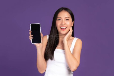 Photo for Beautiful Asian brunette woman cute girl in white tank top ,Excited surprised girl showing smart phone with blank screen , black screen isolated purple background , Mock Up Image - Royalty Free Image