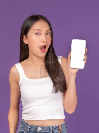 Téléchargez les photos : Portrait de belle jeune femme asiatique montrant le téléphone intelligent avec écran blanc, écran blanc vide espace de copie pour la bannière publicitaire isolé sur fond violet, image maquillée - en image libre de droit