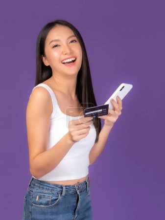 Téléchargez les photos : Belle femme brune asiatique mignonne fille en débardeur blanc tenant carte de crédit et smartphone, téléphone mobile utilise une banque en ligne pour payer des achats en ligne isolé sur fond violet - en image libre de droit