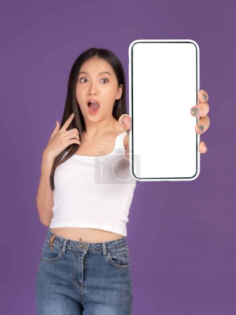 Téléchargez les photos : Beautiful Asian young woman ,Excited surprised girl showing big smart phone with blank screen , white screen isolated on purple background , Display Mock Up Image - en image libre de droit