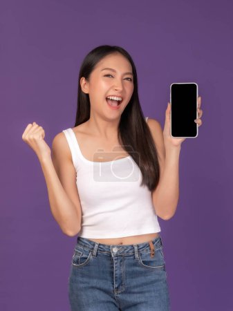 Téléchargez les photos : Portrait de belle jeune femme asiatique montrant l'affichage de téléphone intelligent avec écran vide, écran noir vide espace de copie pour la bannière publicitaire isolé sur fond violet, image maquillée - en image libre de droit