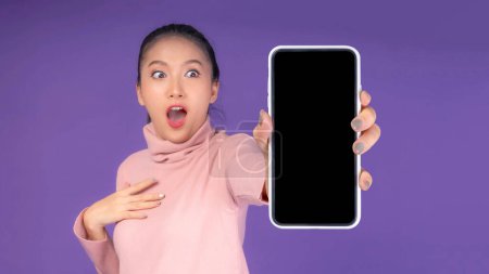 Téléchargez les photos : Belle asiatique jeune femme, excitée surprise fille montrant téléphone intelligent avec écran vide, écran noir isolé sur fond violet, image de maquillage - en image libre de droit