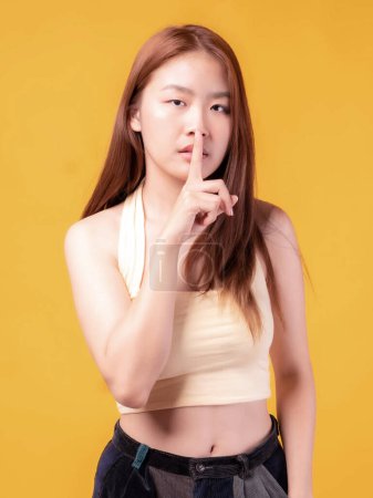 Porträt eines lächelnden eine schöne asiatische junge Frau fühlen sich ernst Schlechte Laune wütend hält seinen Finger in den Mund, macht Schweigegeste isoliert auf gelbem Hintergrund
