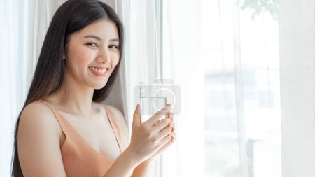 Foto de Hermosa mujer de belleza Asiática linda chica se siente feliz bebiendo agua potable limpia para una buena salud en la mañana, disfrutando del tiempo en su hogar blanco dormitorio fondo estilo de vida belleza mujer concepto - Imagen libre de derechos