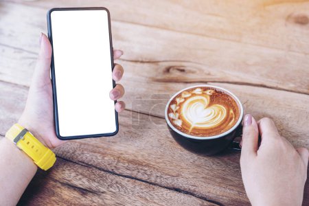 Foto de Taza de café con patrón de corazón de arte latte sobre un fondo de mesa de madera y la mano de la mujer joven sosteniendo el teléfono inteligente en un café cafetería - Imagen libre de derechos