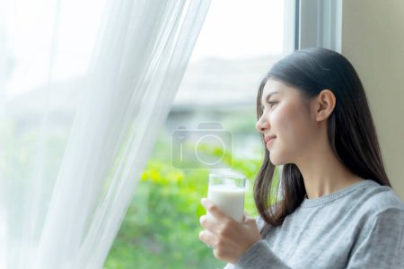 Téléchargez les photos : Belle beauté femme asiatique mignonne fille se sentent heureux boire du lait pour une bonne santé le matin, profiter du temps dans son fond de chambre à coucher blanche à la maison - concept de beauté de style de vie femme - en image libre de droit