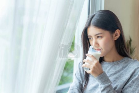 Téléchargez les photos : Belle beauté femme asiatique mignonne fille se sentent heureux boire du lait pour une bonne santé le matin, profiter du temps dans son fond de chambre à coucher blanche à la maison - concept de beauté de style de vie femme - en image libre de droit