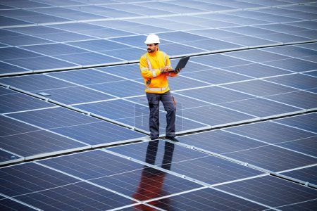 Der Ingenieur für erneuerbare Energien geht durch ein großes Solarkraftwerk und überprüft die Stromerzeugung auf seinem Laptop. Nachhaltige Energiequellen und umweltfreundliche Technologien.