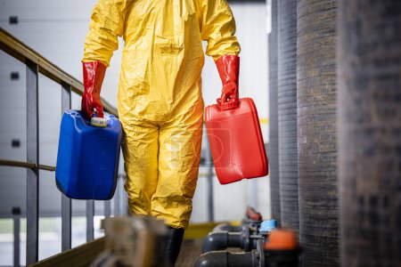 Trabajador irreconocible en traje de protección de materiales peligrosos caminando por depósitos químicos y transportando recipientes de plástico.