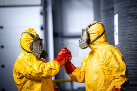 Photo for Factory workers in chemical protection suits and gas masks holding hands together after successful hazardous materials removal. Industrial people teamwork. - Royalty Free Image