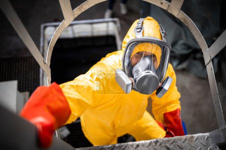 Téléchargez les photos : Travailleur en combinaison de protection et masque à gaz atteignant le haut du réservoir acide à l'intérieur de l'usine de produits chimiques. Risque au travail. Occupation dangereuse. - en image libre de droit