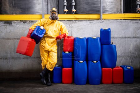 Retrato de trabajador profesional en traje de materiales peligrosos y máscara de gas de pie dentro de la fábrica de productos químicos y la celebración de recipientes.