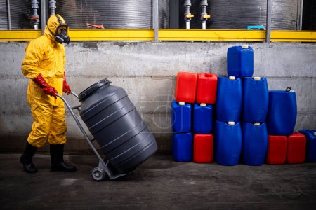 Photo for Professional worker in hazmat suit and gas mask carrying large barrel with acid in chemicals production factory. - Royalty Free Image