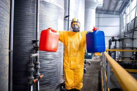 Téléchargez les photos : Portrait d'un ouvrier d'usine expérimenté en combinaison de protection et masque à gaz debout près des réservoirs de stockage d'acide et tenant des conteneurs avec des produits chimiques. - en image libre de droit