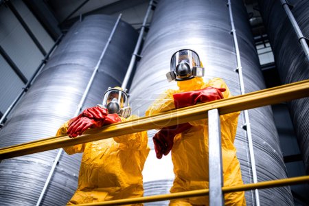 Industrieller Fabrikinnenraum mit Arbeitern in Schutzanzügen, Gasmasken, Handschuhen, die an großen Lagertanks oder Behältern mit Chemikalien stehen.