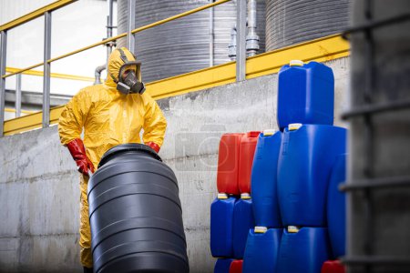 Téléchargez les photos : Travailleur expérimenté en combinaison de protection et masque à gaz déplacer des barils avec de l'acide sulfurique à l'intérieur de l'usine de production de produits chimiques. - en image libre de droit
