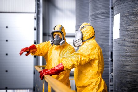 Téléchargez les photos : Factory worker standing by large metal storage tanks with acids wearing yellow protection suit, gas mask and gloves explaining trainee process of chemicals production inside the plant. - en image libre de droit