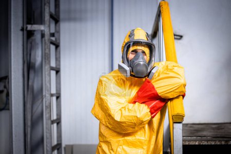 Téléchargez les photos : Portrait d'ouvrier d'usine en combinaison jaune protectrice, masque à gaz et gants appuyés contre la main courante à l'intérieur de l'usine de production de produits chimiques. - en image libre de droit
