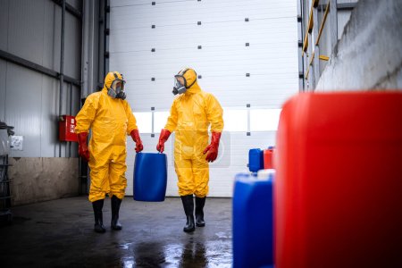 Trabajando en el almacén. Trabajadores en traje amarillo de protección de materiales peligrosos y máscara de gas que transportan recipientes con productos químicos en el almacén.