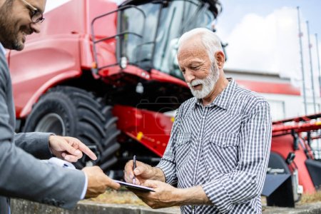 Photo for Senior farmer in plaid shirt signing contract and buying new tractor machine and agricultural equipment at dealership. - Royalty Free Image