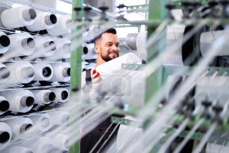 Trabajador de fábrica textil cambiando carrete de hilo en la máquina de tejer industrial.