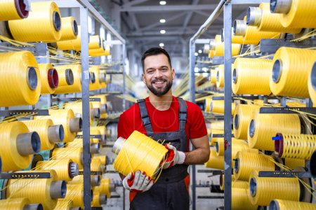 Retrato del trabajador de la fábrica textil sosteniendo carrete de hilo amarillo y de pie por máquina de tejer automatizada.