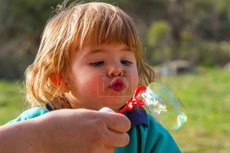 A pretty two-year-old blonde girl plays with blowing soap bubbles on a sunny day