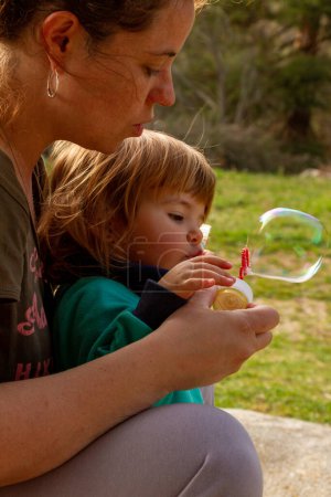 Eine hübsche junge Frau spielt mit ihrer zweijährigen Tochter, um an einem sonnigen Tag Seifenblasen zu pusten