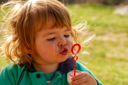 Photo for A pretty two-year-old blonde girl plays with blowing soap bubbles on a sunny day - Royalty Free Image