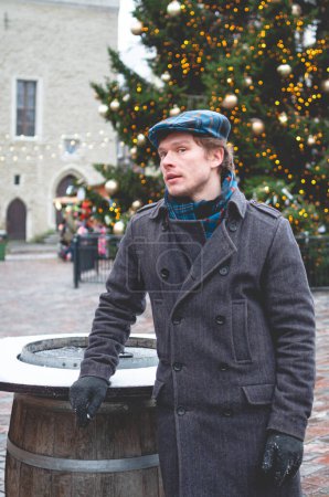 Téléchargez les photos : Un beau jeune homme en manteau gris avec une casquette écossaise en tweed et une écharpe en tartan debout sur une place de l'hôtel de ville de Tallinn par une journée d'hiver lumineuse avec un arbre de Noël et un repas de Noël en arrière-plan. - en image libre de droit