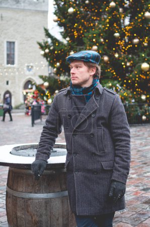Photo for A handsome young man in a grey coat with a Scottish tweed cap and tartan scarf standing on a Tallinn Town Hall Square on a bright winter day with a Christmas tree and a Christmas fare in the background. - Royalty Free Image