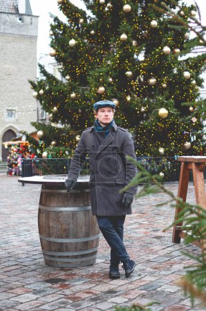 Foto de Un joven guapo con un abrigo gris con una gorra de tweed escocés y una bufanda de tartán soñando mientras está de pie en una plaza del ayuntamiento de Tallin en un brillante día de invierno con un árbol de Navidad y una comida de Navidad en el fondo. - Imagen libre de derechos