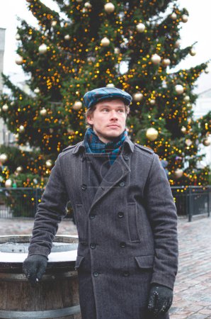 Photo for A handsome young man in a grey coat with a Scottish tweed cap and tartan scarf dreaming while standing on a Tallinn Town Hall Square on a bright winter day with a Christmas tree in the background. - Royalty Free Image