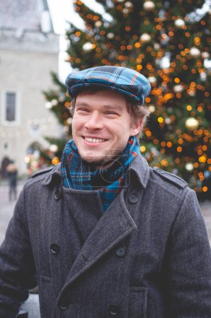 Photo for A handsome young man in a grey coat with a Scottish tweed cap and tartan scarf smiling while standing on a Tallinn Town Hall Square on a bright winter day with a Christmas tree in the background. - Royalty Free Image