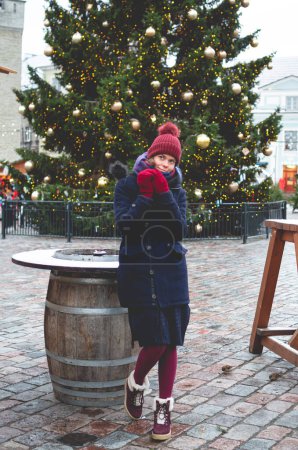 Photo for A young handsome smiling woman dreaming while holding hands together and standing on a Tallinn Town Hall Square on a cold winter day with a Christmas tree in the background. - Royalty Free Image