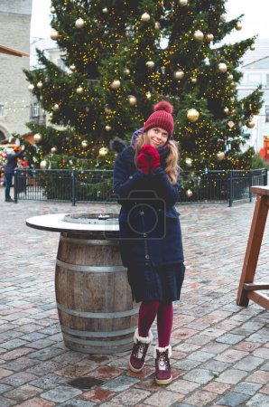 Photo for A young handsome smiling woman dreaming while holding hands together and standing on a Tallinn Town Hall Square on a cold winter day with a Christmas tree in the background. - Royalty Free Image
