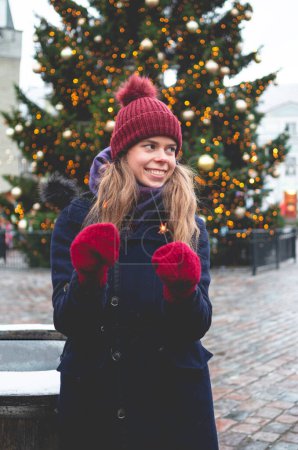 Photo for A young handsome smiling woman holding sparklers and standing on a Tallinn Town Hall Square on a cold winter day with a Christmas tree in the background. - Royalty Free Image