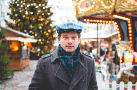 Photo for A handsome young man in a grey coat with a Scottish tweed cap and tartan scarf standing on a Tallinn Town Hall Square on a bright winter day with a Christmas tree and a Christmas fare in the background. - Royalty Free Image